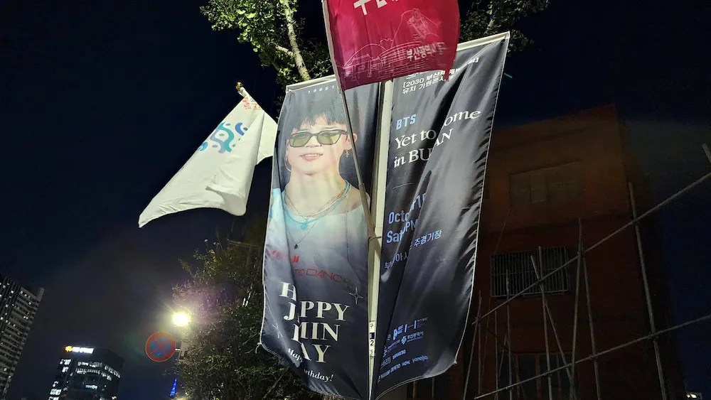 Night view of a banner saying “HBD Jimin” near Cafe Magnate, glowing under streetlights