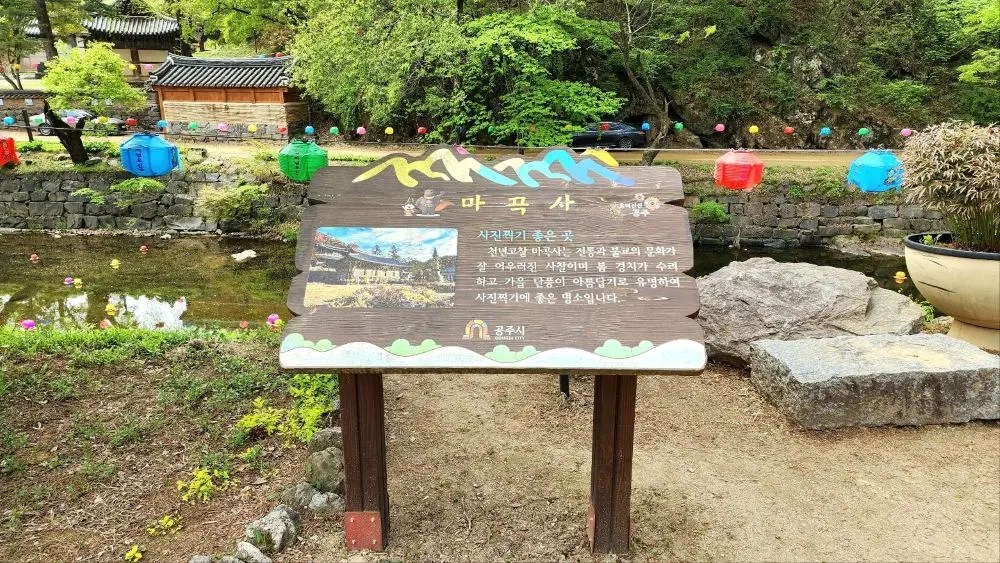 An information sign describing Magoksa Temple in Gongju, South Korea, placed beside a small stream decorated with colorful lanterns.