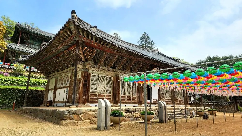 A traditional wooden hall at Magoksa Temple in Gongju, South Korea, with intricate roof architecture and rows of colorful lanterns displayed in the courtyard.