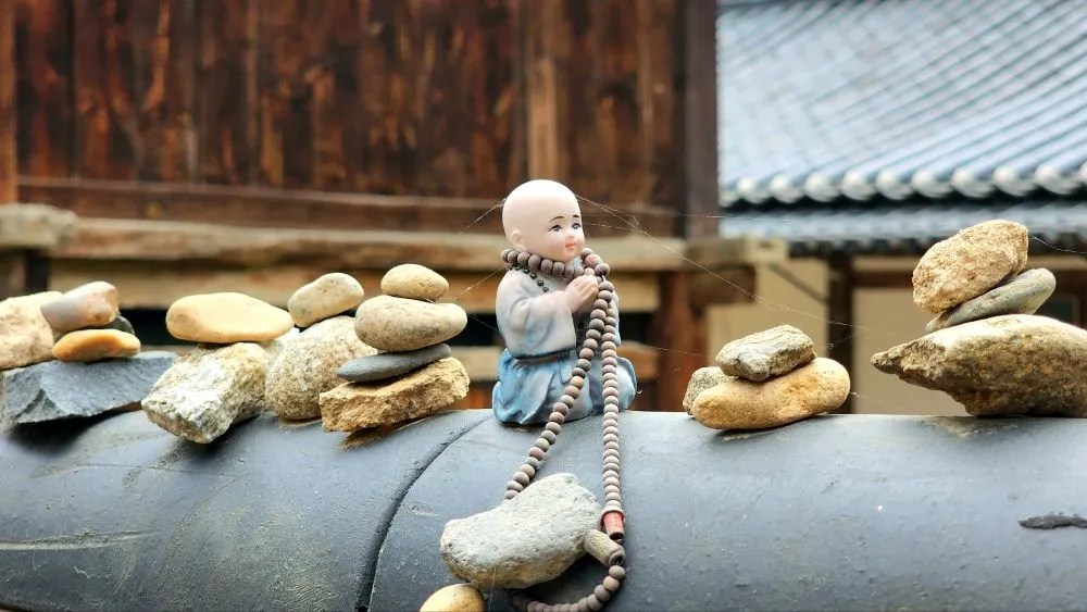 A close-up of a small monk figurine holding prayer beads, placed among stacks of stones at Magoksa Temple in Gongju, South Korea.