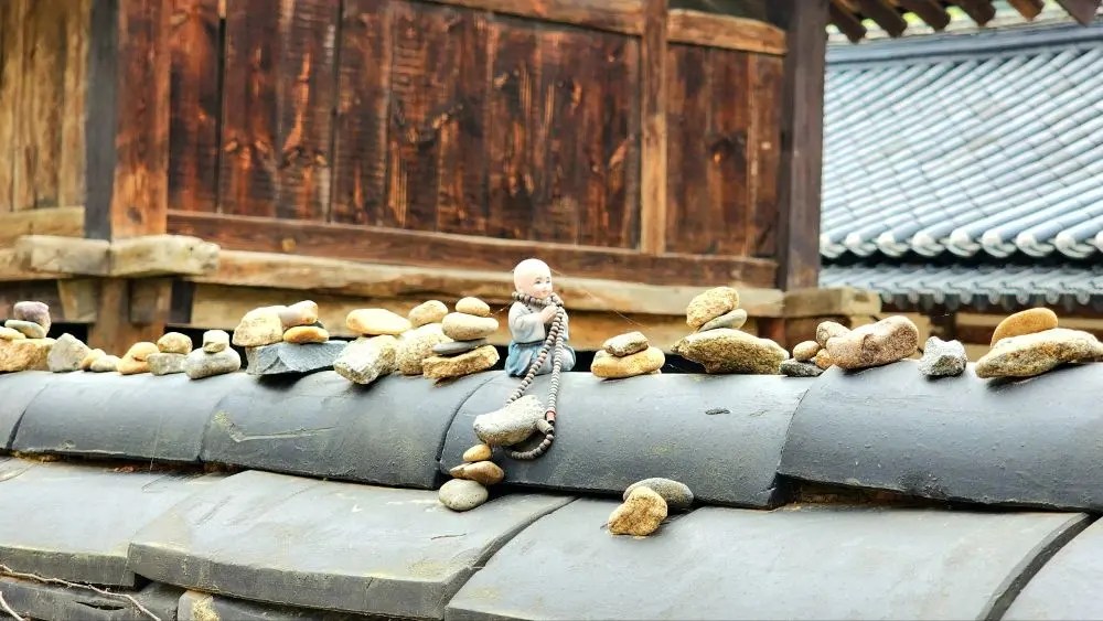 A small monk figurine holding prayer beads placed among stacked stones on a temple wall at Magoksa Temple in Gongju, South Korea.