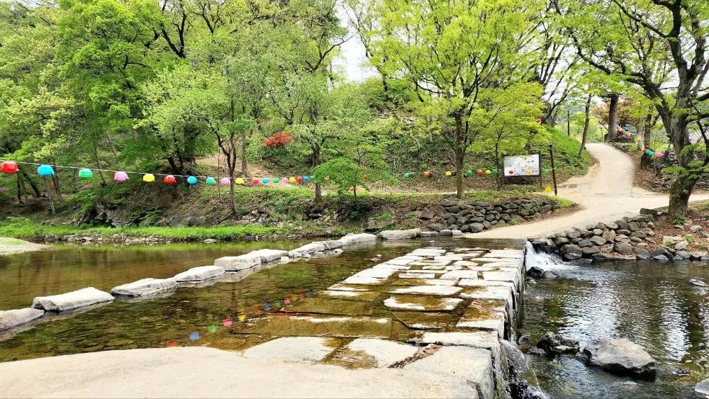 Stepping stones crossing a calm stream at Magoksa Temple in Gongju, South Korea, with a line of colorful lanterns hanging among fresh spring trees.