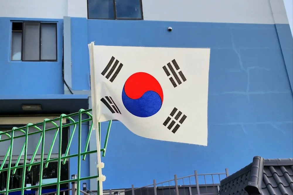 Drapeau coréen au premier plan avec en arrière-plan un bâtiment bleu et une clôture verte / Korean flag in the foreground with a blue building and green fence in the background