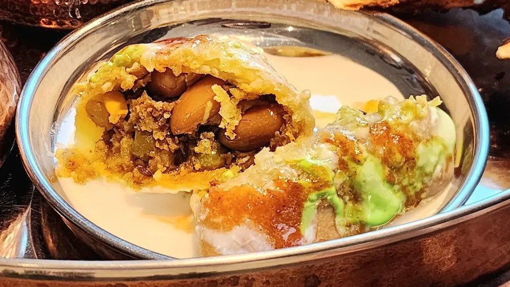 A thali with samosa topped with yogurt and sauces, naan, and various Indian dishes on a tray.