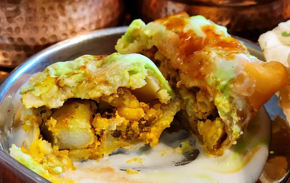 A thali with samosa topped with yogurt and sauces, naan, and various Indian dishes on a tray.