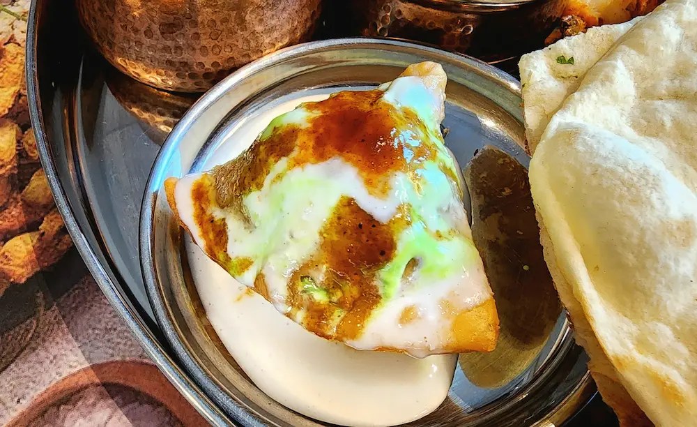A thali with samosa topped with yogurt and sauces, naan, and various Indian dishes on a tray.