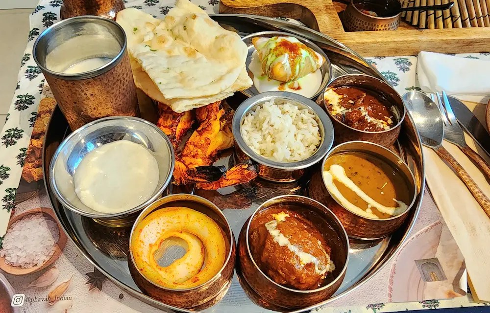 South Indian thali with various curries, naan, rice, tandoori shrimp, and chutneys — served on a round metal tray.