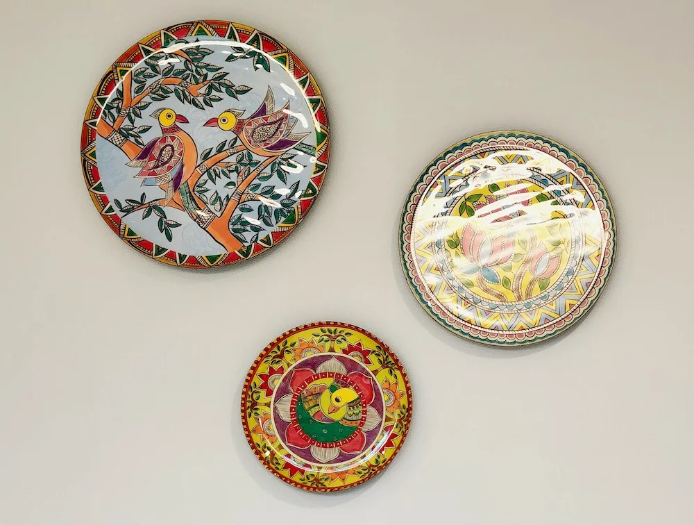 Three colorful decorative plates with Indian folk designs — birds, trees, and floral patterns on a neutral wall.
