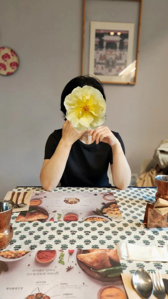 My aunt sitting at Bhavan restaurant in Busan, with her face covered by a flower sticker. A soft light falls on the table.