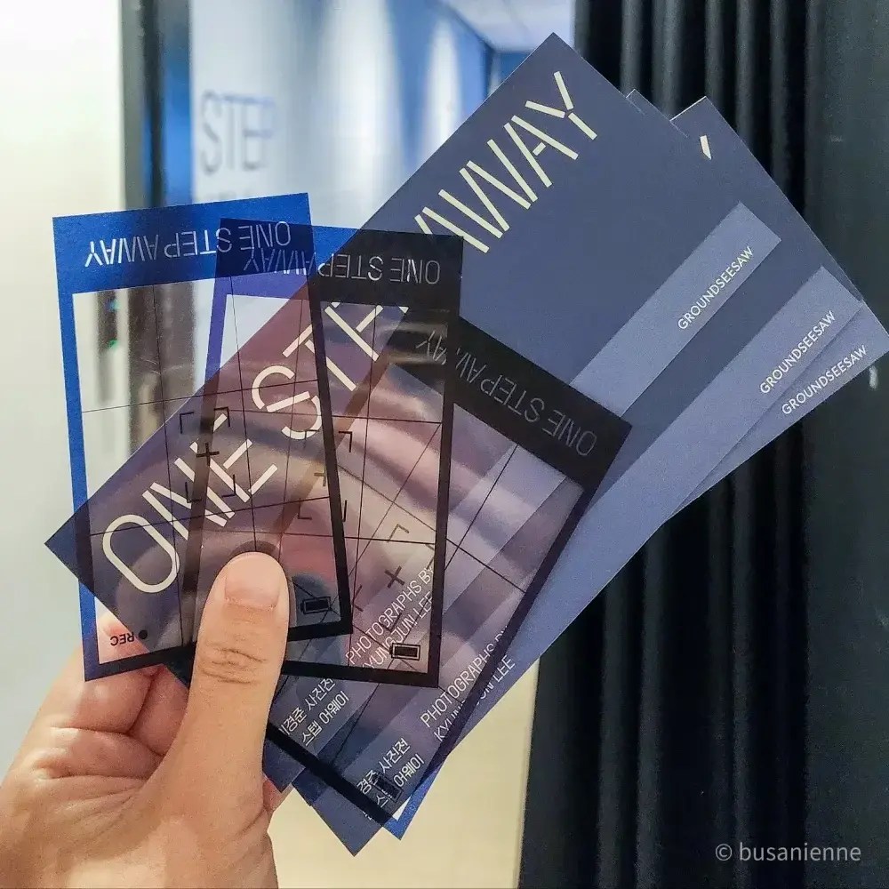 Main tenant des billets et cartes transparentes offertes à l’exposition photo « One Step Away » de Kyungjun Lee à Busan