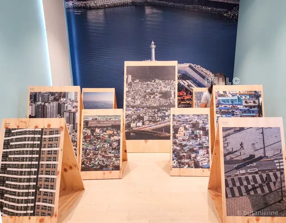 Photographies de Busan exposées sur des panneaux en bois avec un phare en arrière-plan, une visiteuse visible partiellement