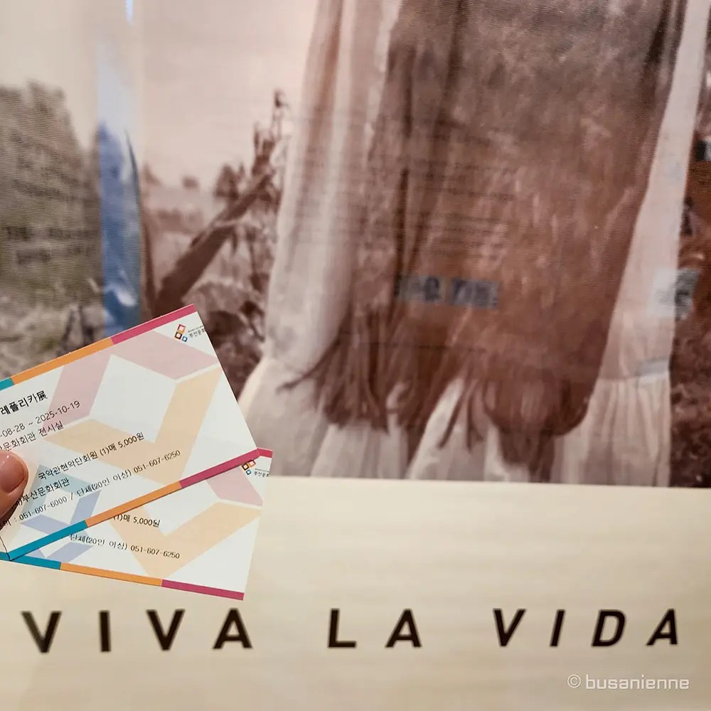 Two colorful exhibition tickets held over a white wall with the large phrase 'VIVA LA VIDA' and a faded photograph of a woman in the background. / Deux billets d'exposition colorés tenus au-dessus d'un mur blanc avec la grande phrase 'VIVA LA VIDA' et une photo estompée d'une femme en arrière-plan.