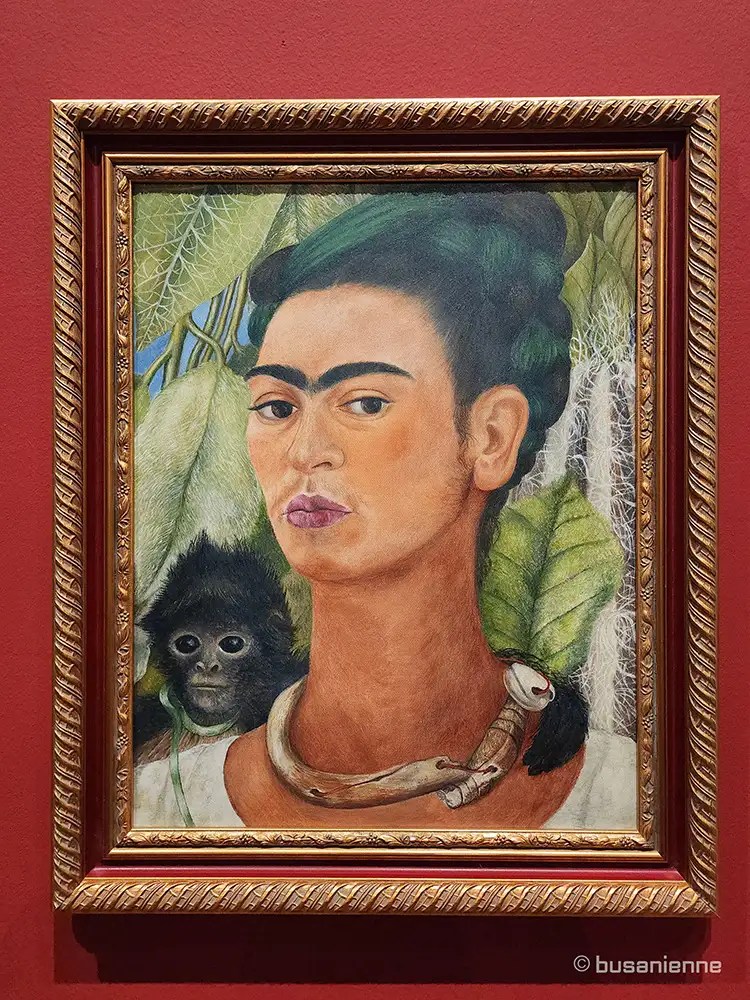 Replica of Frida Kahlo's 'Self-Portrait with Monkey' (1938). The artist stares intensely at the viewer, wearing a bone necklace and a purple head wrap with butterflies. A spider monkey sits on her left shoulder, and a black cat is behind her right shoulder, all surrounded by large green leaves.