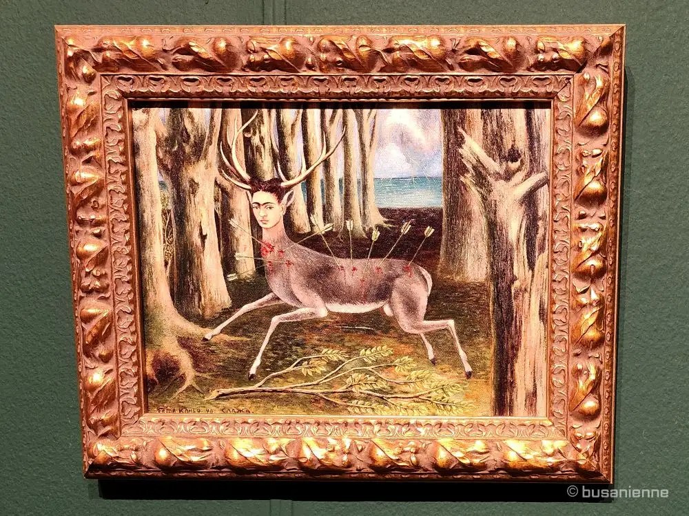 Replica of Frida Kahlo's painting 'The Wounded Deer' (El venado herido), a self-portrait where she appears as a deer pierced with arrows, running through a forest. The artwork is displayed in an ornate gold frame at the Busan Exhibition.
