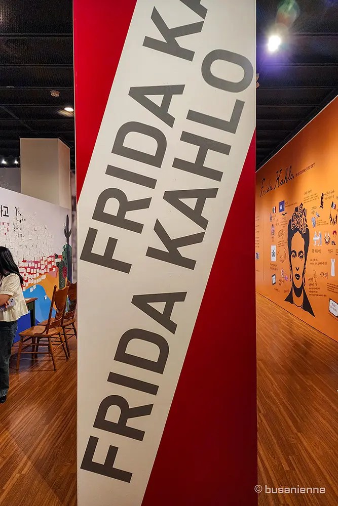 A white pillar with a red diagonal stripe featuring the large, stacked name 'FRIDA KAHLO' in black capital letters inside the exhibition hall. The orange biography wall is visible in the background. / Un pilier blanc avec une bande diagonale rouge affichant le nom empilé 'FRIDA KAHLO' en grandes lettres noires à l'intérieur de la salle d'exposition. Le mur biographique orange est visible en arrière-plan.