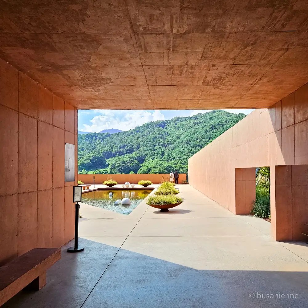 Red concrete walls leading to a reflecting pool with spherical sculptures and mountain backdrop at Sihaon Café in Ulsan.