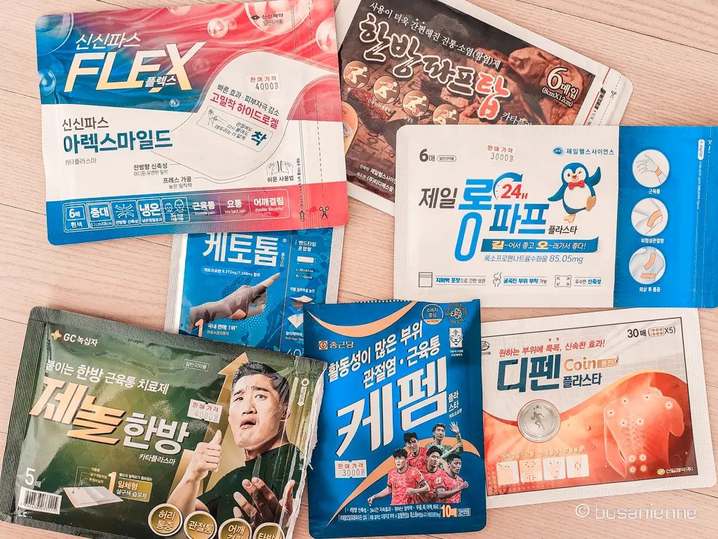 A tabletop flatlay featuring different Korean pain-relief patches from multiple brands, highlighting various cooling, heating, and herbal formulas.