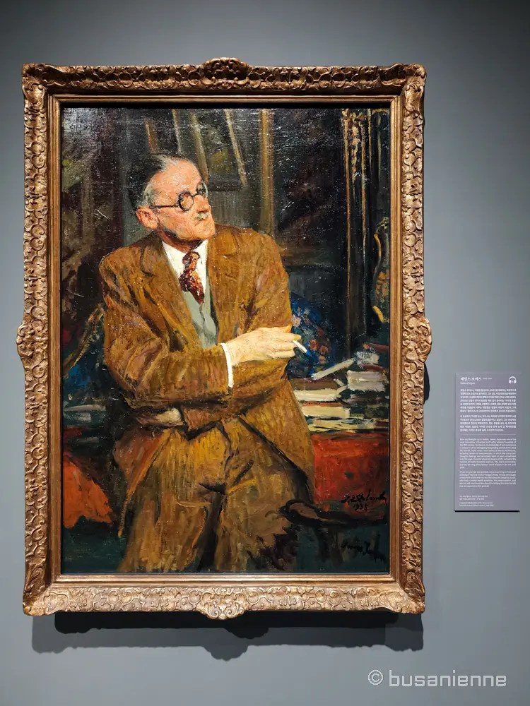 Portrait of Irish writer James Joyce seated indoors, wearing round glasses and a brown suit, painted by Jacques-Émile Blanche in 1935, with books and papers behind him