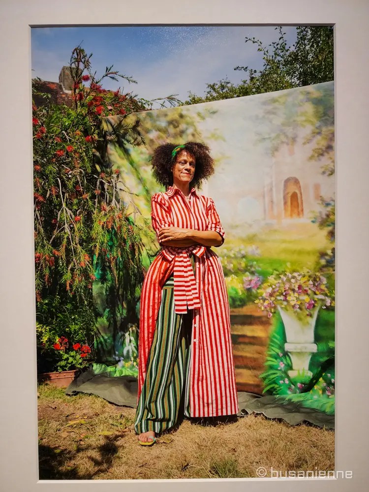 Portrait of British writer Bernardine Evaristo standing outdoors, photographed by Ruth Ossai in 2020, wearing a red striped dress in a staged garden setting
